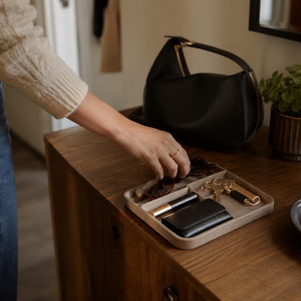 A hand reaches into an entryway tray holding keys, a wallet, and cards beside a handbag.